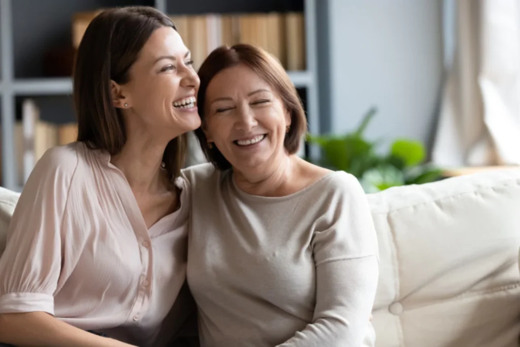 Two women, mother and adult daughter sitting on a white couch with arm around each other, smiling
