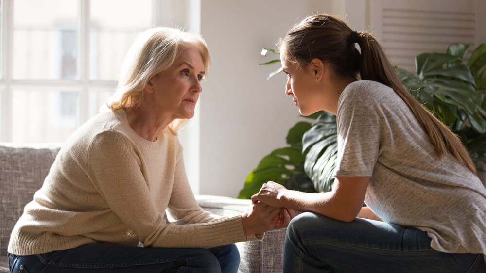 Mom and young adult daughter sitting, holding hands, and having heart-to-heart talk