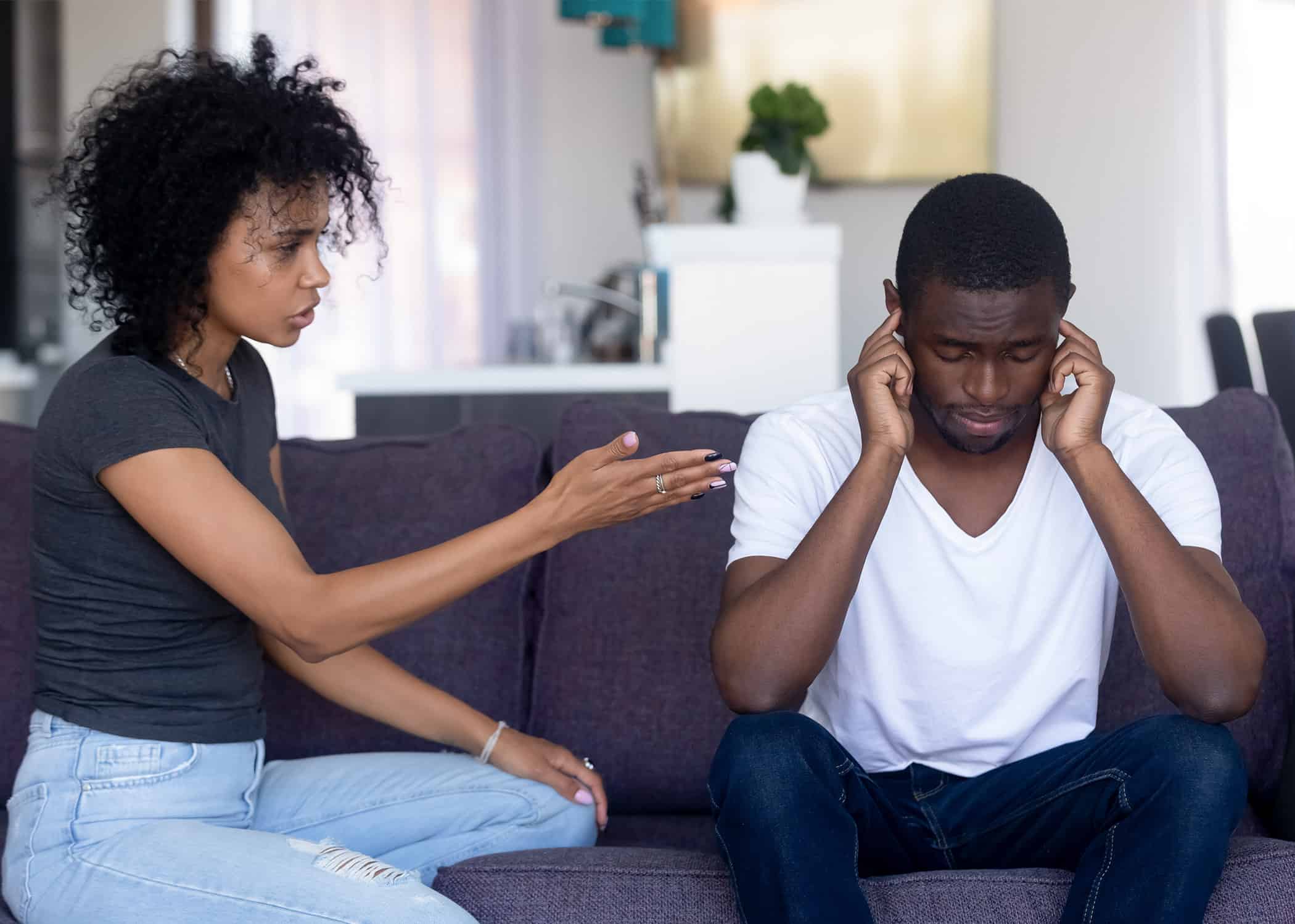 Controlling-black-wife-lecturing-her-black-husband-sitting-on-couch-with-hands-over-ears